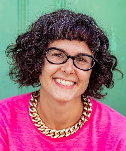 A picture of a smiling white woman with brown curly hair, wearing black framed glasses, a pink t. shirt and a gold, chain necklace.