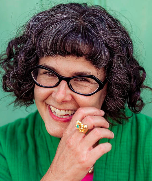 A close up of a smiling white woman with curly brown hair wearing black framed glasses and a green jacket. Her hand is near her face and she is wearing a gold and green ring.