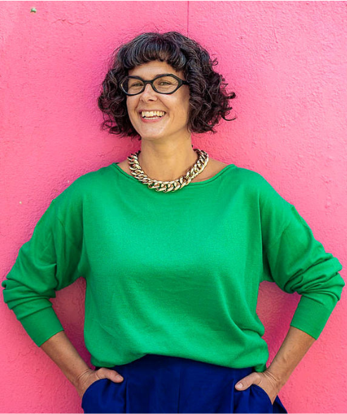 A smiling white woman with curly brown hair, wearing a green jumper and a gold necklace, standing against a pink wall.