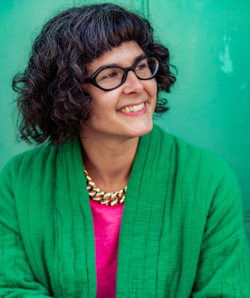 A smiling white woman wearing black framed glasses, a green jacket and pink t.shirt with a gold, chain necklace, sitting against a green wall